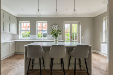 This is an example of a large classic grey and white u-shaped kitchen/diner in London with an integrated sink, shaker cabinets, grey cabinets, quartz worktops, white splashback, engineered quartz splashback, black appliances, light hardwood flooring, an island, brown floors, white worktops and feature lighting.