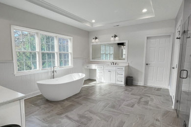 Photo of a large transitional master bathroom in Philadelphia with recessed-panel cabinets, white cabinets, a freestanding tub, a double shower, a one-piece toilet, gray tile, ceramic tile, grey walls, porcelain floors, an undermount sink, engineered quartz benchtops, grey floor, a hinged shower door, white benchtops, a niche, a double vanity, a built-in vanity, recessed and decorative wall panelling.
