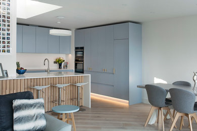 Mid-sized scandinavian l-shaped eat-in kitchen in Other with louvered cabinets, blue cabinets, quartzite benchtops, white splashback, stainless steel appliances, light hardwood floors, with island and white benchtop.