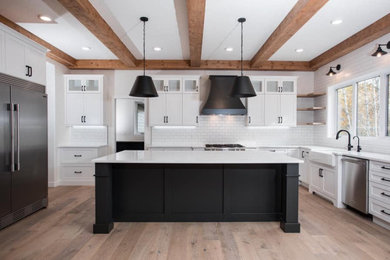 Large country u-shaped eat-in kitchen in Edmonton with a farmhouse sink, shaker cabinets, black cabinets, quartz benchtops, white splashback, ceramic splashback, stainless steel appliances, light hardwood floors, with island, brown floor and white benchtop.