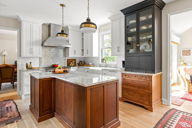 Example of a mid-sized classic l-shaped light wood floor and beige floor enclosed kitchen design in Philadelphia with a farmhouse sink, recessed-panel cabinets, granite countertops, white backsplash, ceramic backsplash, stainless steel appliances, an island and multicolored countertops