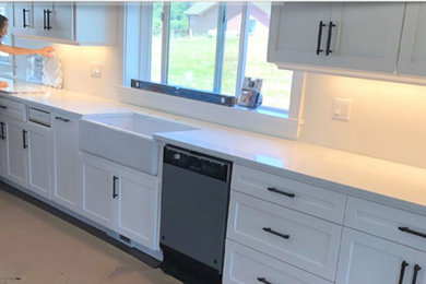 This is an example of a medium sized contemporary l-shaped kitchen/diner in Portland with a belfast sink, shaker cabinets, white cabinets, engineered stone countertops, white splashback, ceramic splashback, stainless steel appliances, light hardwood flooring, an island, beige floors, white worktops and a coffered ceiling.