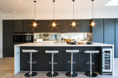 Inspiration for a large modern single-wall open plan kitchen in Surrey with a built-in sink, flat-panel cabinets, grey cabinets, composite countertops, metallic splashback, glass sheet splashback, black appliances, ceramic flooring, an island and grey floors.