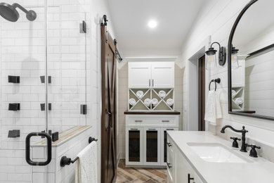 Mid-sized country master white tile and subway tile painted wood floor, brown floor, double-sink and shiplap wall alcove shower photo in Milwaukee with shaker cabinets, white cabinets, white walls, an undermount sink, quartz countertops, a hinged shower door, gray countertops, a niche and a built-in vanity