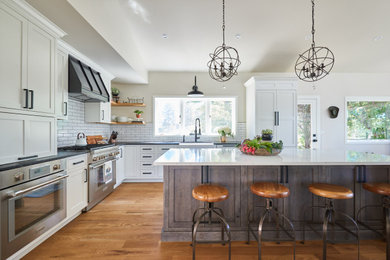 Photo of a large country l-shaped open plan kitchen in Denver with a farmhouse sink, shaker cabinets, white cabinets, quartz benchtops, white splashback, porcelain splashback, stainless steel appliances, medium hardwood floors, with island, brown floor and black benchtop.