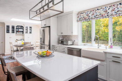 Large elegant l-shaped medium tone wood floor and brown floor eat-in kitchen photo in Boston with a farmhouse sink, recessed-panel cabinets, white cabinets, quartz countertops, gray backsplash, ceramic backsplash, stainless steel appliances, an island and white countertops