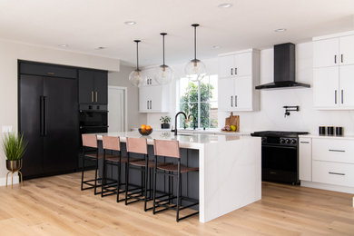 Example of a trendy light wood floor and brown floor eat-in kitchen design in Orange County with a drop-in sink, flat-panel cabinets, white cabinets, quartzite countertops, white backsplash, quartz backsplash, black appliances, an island and white countertops