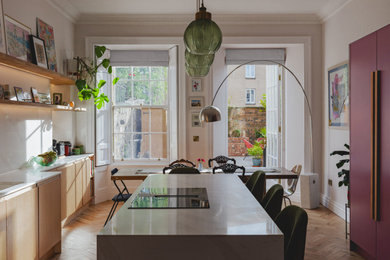 Example of a mid-sized trendy galley medium tone wood floor eat-in kitchen design in Edinburgh with a drop-in sink, flat-panel cabinets, light wood cabinets, marble countertops, white backsplash, marble backsplash, paneled appliances, an island and white countertops