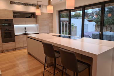Large contemporary kitchen/diner in London with a single-bowl sink, flat-panel cabinets, grey cabinets, engineered stone countertops, white splashback, engineered quartz splashback, integrated appliances, light hardwood flooring, an island, beige floors and grey worktops.