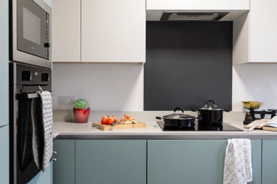 Bild på ett avskilt, litet funkis vit vitt l-kök, med en nedsänkt diskho, släta luckor, gröna skåp, bänkskiva i koppar, grått stänkskydd, glaspanel som stänkskydd och rostfria vitvaror