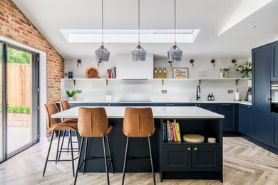 Inspiration for a large classic l-shaped open plan kitchen in Surrey with a submerged sink, shaker cabinets, blue cabinets, quartz worktops, white splashback, engineered quartz splashback, black appliances, medium hardwood flooring, an island, brown floors, white worktops, a vaulted ceiling and a feature wall.