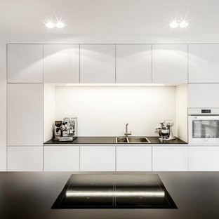 Offene Moderne Küche mit Doppelwaschbecken, flächenbündigen Schrankfronten, weißen Schränken, Küchenrückwand in Weiß, Kücheninsel, weißen Elektrogeräten und Betonboden in Köln