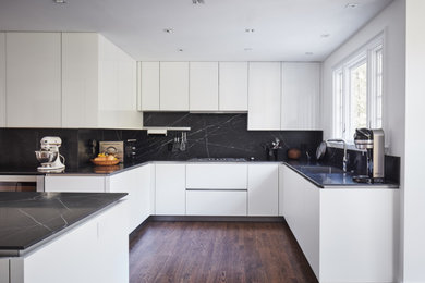 This is an example of a large modern eat-in kitchen in New York with an undermount sink, glass-front cabinets, white cabinets, black splashback, stainless steel appliances, dark hardwood floors, with island, brown floor and black benchtop.