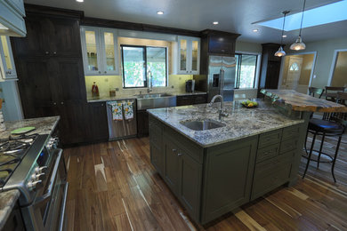 Example of a large transitional l-shaped medium tone wood floor and brown floor eat-in kitchen design in Sacramento with a farmhouse sink, shaker cabinets, dark wood cabinets, granite countertops, beige backsplash, travertine backsplash, stainless steel appliances, an island and multicolored countertops