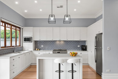 This is an example of a mid-sized transitional u-shaped open plan kitchen in Sydney with an undermount sink, shaker cabinets, white cabinets, quartz benchtops, grey splashback, ceramic splashback, stainless steel appliances, light hardwood floors, with island, brown floor and grey benchtop.