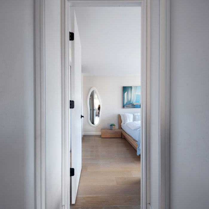 This serene modern bedroom is revealed through a clean, minimalist hallway framed by crisp white walls and sleek black hardware. The space features light oak hardwood floors, creating a warm, natural