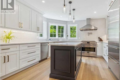 Photo of a large contemporary u-shaped separate kitchen in Toronto with an undermount sink, shaker cabinets, white cabinets, quartz benchtops, white splashback, ceramic splashback, stainless steel appliances, light hardwood floors, with island, brown floor and white benchtop.