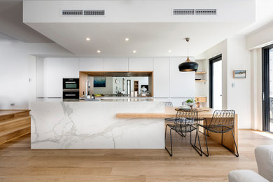 Photo of a large scandi galley open plan kitchen in Perth with a single-bowl sink, flat-panel cabinets, white cabinets, engineered stone countertops, metallic splashback, mirror splashback, black appliances, light hardwood flooring, an island, brown floors and white worktops.