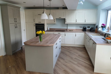 Photo of a medium sized contemporary l-shaped kitchen/diner in West Midlands with a double-bowl sink, shaker cabinets, beige cabinets, laminate countertops, black appliances, laminate floors, an island, brown floors, brown worktops and a coffered ceiling.
