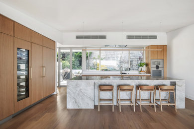This is an example of a large beach style l-shaped open plan kitchen in Sydney with a single-bowl sink, recessed-panel cabinets, medium wood cabinets, quartzite benchtops, white splashback, engineered quartz splashback, stainless steel appliances, medium hardwood floors, with island, brown floor, grey benchtop and vaulted.