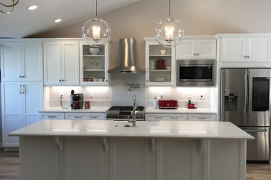 Mid-sized minimalist single-wall light wood floor and brown floor eat-in kitchen photo in Other with a farmhouse sink, white cabinets, granite countertops, white backsplash, stainless steel appliances, an island and white countertops