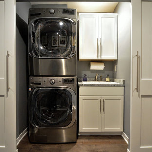 Design ideas for a small traditional single-wall laundry cupboard in Minneapolis with a single-bowl sink, white cabinets, grey walls, medium hardwood flooring, a stacked washer and dryer, brown floors and shaker cabinets.