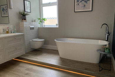 Inspiration for a large contemporary grey and white family bathroom in Gloucestershire with shaker cabinets, grey cabinets, a freestanding bath, a walk-in shower, a wall mounted toilet, grey walls, wood-effect flooring, a wall-mounted sink, brown floors, an open shower, white worktops, feature lighting, double sinks and a floating vanity unit.