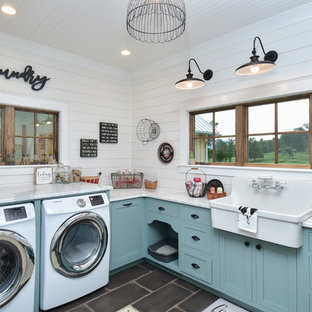 Modelo de lavadero multiusos y en U, campestre, con fregadero sobremueble, armarios estilo shaker, puertas de armario azules, encimera de mármol, paredes blancas, suelo de baldosas de porcelana, lavadora y secadora integrada, suelo negro y encimeras blancas
