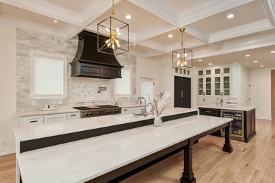 Klassisk inredning av ett mycket stort vit vitt kök och matrum, med en rustik diskho, luckor med infälld panel, vita skåp, bänkskiva i kvarts, grått stänkskydd, stänkskydd i marmor, svarta vitvaror, ljust trägolv, flera köksöar och brunt golv