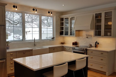 Photo of a mid-sized transitional l-shaped eat-in kitchen in Ottawa with a farmhouse sink, shaker cabinets, beige cabinets, quartz benchtops, white splashback, ceramic splashback, stainless steel appliances, light hardwood floors, with island, yellow floor and white benchtop.