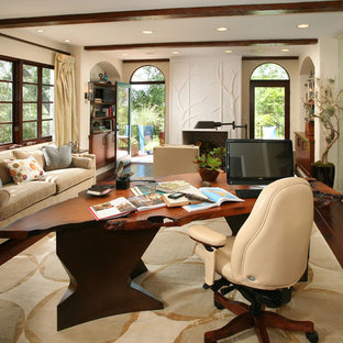 This is an example of a large transitional study room in Los Angeles with dark hardwood floors, a freestanding desk, beige walls, a standard fireplace, a plaster fireplace surround and brown floor.