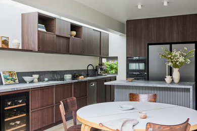 This is an example of a mid-sized midcentury l-shaped eat-in kitchen in Sydney with an undermount sink, flat-panel cabinets, dark wood cabinets, quartz benchtops, grey splashback, engineered quartz splashback, black appliances, travertine floors, with island, beige floor and grey benchtop.