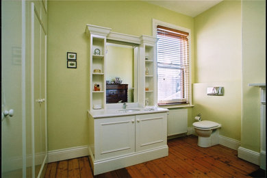 Modelo de cuarto de baño único y a medida clásico pequeño con armarios tipo mueble, puertas de armario blancas, sanitario de pared, paredes verdes, suelo de madera clara, aseo y ducha, encimera de granito, suelo marrón y encimeras blancas