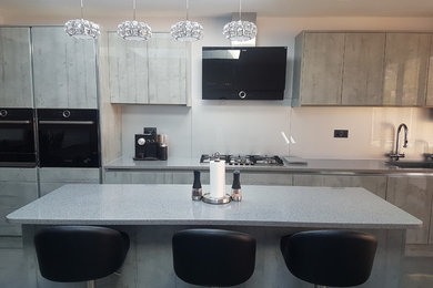 This is an example of a medium sized contemporary galley open plan kitchen in London with a submerged sink, flat-panel cabinets, medium wood cabinets, quartz worktops, grey splashback, glass sheet splashback, black appliances, an island and grey worktops.