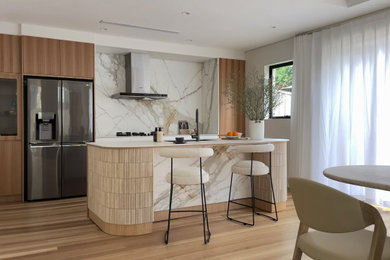 This is an example of a mid-sized contemporary single-wall eat-in kitchen in Sydney with a double-bowl sink, flat-panel cabinets, medium wood cabinets, solid surface benchtops, white splashback, marble splashback, black appliances, medium hardwood floors, with island and white benchtop.