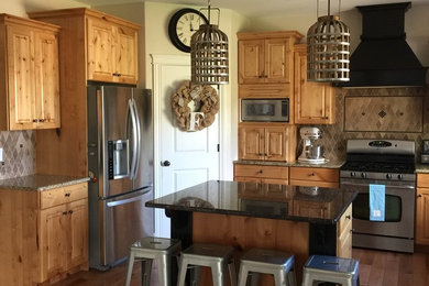 Example of a mid-sized mountain style l-shaped dark wood floor and brown floor kitchen design in Austin with raised-panel cabinets, medium tone wood cabinets, granite countertops, brown backsplash, ceramic backsplash, stainless steel appliances and an island