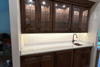 Inspiration for a small timeless single-wall medium tone wood floor and brown floor wet bar remodel in Cleveland with an undermount sink, glass-front cabinets, dark wood cabinets, quartzite countertops, white backsplash and white countertops