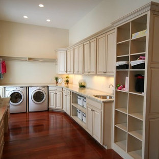 クリーブランドの大きい地中海スタイルのおしゃれな家事室 (アンダーカウンターシンク、レイズドパネル扉のキャビネット、ベージュのキャビネット、御影石カウンター、ベージュの壁、濃色無垢フローリング、左右配置の洗濯機・乾燥機、赤い床) の写真