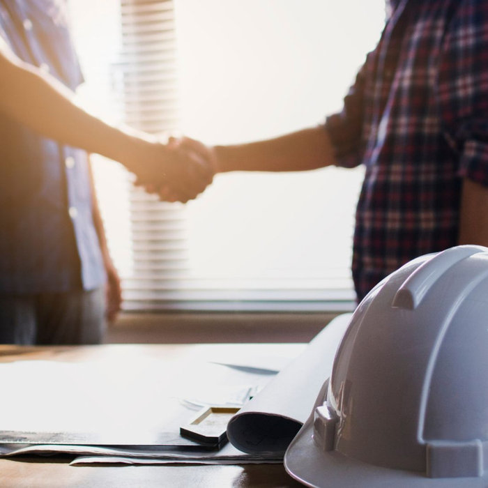 Two people shaking hands across a table with a hard hat, symbolizing trust and partnership in home remodeling.