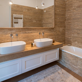 Mid-sized trendy master beige tile and porcelain tile light wood floor and yellow floor drop-in bathtub photo in Milan with recessed-panel cabinets, white cabinets, a wall-mount toilet, white walls, a vessel sink, tile countertops and beige countertops