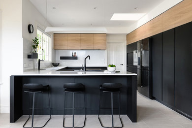 Photo of a contemporary u-shaped separate kitchen in Hobart with a drop-in sink, flat-panel cabinets, black cabinets, white splashback, black appliances, a peninsula, beige floor and white benchtop.