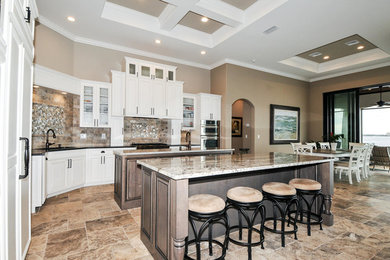Example of a mid-sized tuscan single-wall travertine floor eat-in kitchen design in Miami with a single-bowl sink, raised-panel cabinets, white cabinets, granite countertops, multicolored backsplash, ceramic backsplash, stainless steel appliances and two islands