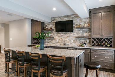 Large transitional single-wall brown floor seated home bar photo in DC Metro with brown cabinets, granite countertops, beige backsplash, stone tile backsplash, recessed-panel cabinets, an undermount sink and gray countertops