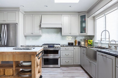 Example of a trendy l-shaped light wood floor kitchen design in Cincinnati with a farmhouse sink, recessed-panel cabinets, beige cabinets, white backsplash, glass tile backsplash, stainless steel appliances and an island