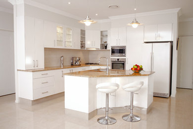 This is an example of a mid-sized contemporary l-shaped eat-in kitchen in Brisbane with an undermount sink, flat-panel cabinets, white cabinets, granite benchtops, white splashback, cement tile splashback, stainless steel appliances, ceramic floors, with island and beige floor.