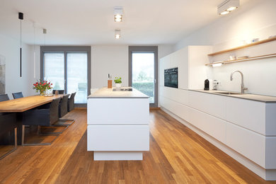 Photo of an expansive contemporary galley open plan kitchen in Dusseldorf with an undermount sink, flat-panel cabinets, white cabinets, quartz benchtops, white splashback, glass sheet splashback, stainless steel appliances, painted wood floors, with island, brown floor and grey benchtop.