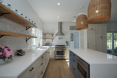 Inspiration for a beach style l-shaped kitchen in Boston with an undermount sink, shaker cabinets, white cabinets, solid surface benchtops, white splashback, subway tile splashback, stainless steel appliances, light hardwood floors, with island, beige floor and white benchtop.