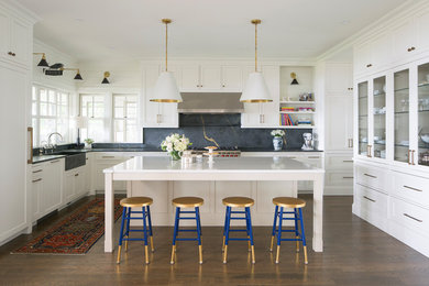 Beach style u-shaped dark wood floor and brown floor kitchen photo in Minneapolis with a farmhouse sink, shaker cabinets, white cabinets, gray backsplash, stone slab backsplash, stainless steel appliances and an island