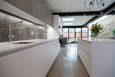 Photo of a medium sized contemporary galley open plan kitchen in London with a submerged sink, flat-panel cabinets, white cabinets, composite countertops, grey splashback, porcelain splashback, black appliances, concrete flooring, an island, grey floors and grey worktops.