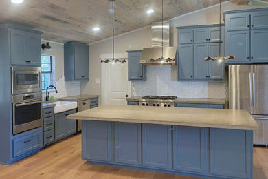 Medium tone wood floor kitchen photo with a farmhouse sink, shaker cabinets, blue cabinets, concrete countertops, white backsplash, porcelain backsplash, stainless steel appliances and an island